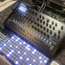 Černý syntezátor Roland SYNTHCUBER SH-101 s displejem, faderem a řadou otočných knoflíků. Vedle něj se nachází černý kontrolér značky OX s mřížkou mo…