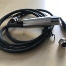 Na fotografii je černý kabel s kovovým cylindrickým koncem, který má v horní části svislé rýhy. Na kabelu je viditelný bílý nápis SHURE. Součástí jso…