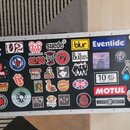Černý transportní kufr s hliníkovými hranami a kovovými rohy, který je polepen mnoha nálepkami. Mezi viditelnými nápisy a logy jsou: Apple, U2, RADIO…