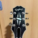 Hlava kytary v černé barvě s bílým lemováním. V horní části je bílý nápis Epiphone a uprostřed se nachází vložka ve tvaru kříže z perleťového materiá…