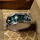 Zelený snare drum s kovovými obručmi a mechanismem na napnutí blány. Na bílé bláně je viditelné logo značky Ludwig. Povrch nástroje má texturovaný, v…