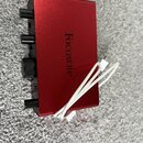 Červený kovový audio interface s nápisem Focusrite na horní straně. Na přední hraně jsou vidět dva černé oválné knofíky a jeden větší kulatý regulačn…