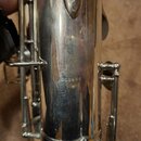 Detailní záběr na kovový, stříbrný povrch dechového nástroje s mechanikou. Na těle nástroje je vyryto sériové číslo 262498. Povrch vykazuje drobné šk…