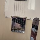 Detail elektrické kytary v krémové barvě s bílým pickguardem. Na chromovaném krytu mostu je vidět nápis FENDER a černý humbucker. Kytara má kovovou o…