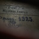 Detailní záběr na vnitřní štítkování smyčcového nástroje z dřeva. Je zde viditelný tištěný nápis Nicolaus Amatus a pod ním ručně psaný text v modrém…
