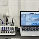 Fotografie zachicuje pracovní stůl s hudební technikou. Vlevo je bílá rytmická sekvenční jednotka s nápisem Roland TR-707, umístěná na černém studiov…