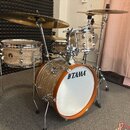 Akustická bicí souprava s povrchem v imitaci světlého dřeva. Na basovém bubnu je viditelné logo TAMA. Souprava obsahuje několik činelů, tom a snare d…