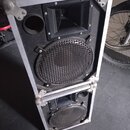 Dvě černé reproduktorové bedny v hliníkových transportních kufrech s kovovými rohy. Horní bedna obsahuje jeden velký reproduktor s mřížkou a jeden vý…