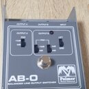 Šedý kovový přístroj s nápisem AB-O BALANCED LINE OUTPUT SWITCHER a logem Palmer Musical Instruments. Na horní straně jsou tři přepínače s popisky PA…
