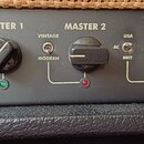 Detail ovládacího panelu kytarového zesilovače v černé barvě. Jsou zde viditelné popisky MASTER 1, MASTER 2, TREBLE a MIDDLE spolu s odpovídajícími o…