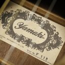 Fotografie detailu vnitřní štítku akustické kytary. Na štítku je vidět ornamentální rám s nápisem Yamaki a texty YAMAKI MUSICAL INSTRUMENTS CO.,LTD.…