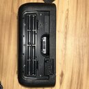 Fotografie zachycuje zadní stranu černého elektronického zařízení s texturovaným povrchem. Je vidět otevřený prostor pro baterie, D-sub konektor a vs…