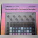 Fotografie zobrazuje sampler Novation Circuit Rhythm v originálním fialovém balení. Zařízení má šedý povrch, osm ovládacích knoflíků a mřížku podsvíc…