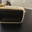 Bílý přenosný reproduktor s povrchem imitujícím kůži a zlatým ovládacím knoflíkem. Zadní strana je opatřena černým mřížkovým krytem, v jehož středě j…