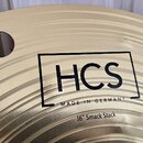 Detailní záběr na zlatavý kovový činel s koncentrickými rýhami. Uprostřed je černý obdélníkový rám s nápisem HCS a pod ním text MADE IN GERMANY. Dole…