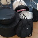 Sada černých přepravních tašek různých velikostí, z nichž dvě mají bílý nápis Millenium a jedna nápis Groove. V pozadí je vidět bílý bicí komplet s n…