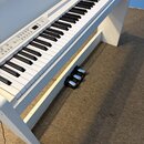 Bílé digitální piano s klaviatura v bílo-černém provedení. Na ovládacím panelu jsou vidět tlačítka a přepínače, ale nápisy nejsou čitelné. Pod klavia…