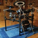 Elektronická bicí souprava značky Roland na černém kovovém rámu, umístěná na modrém koberci. Na basovém bubnu je bílý nápis Roland a v dolní části lo…