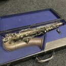 V modrém polstrování kufru leží saxofon s matným, stříbrno-šedým povrchem. Nástroj vykazuje známky opotřebení a oxidace materiálu, zejména v oblasti…