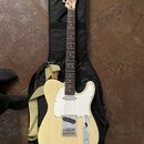 Krémová elektrická kytara typu Telecaster s bílým pickguardem, položená na černém softcasu. Na hlavě hředu je viditelné logo Fender. Kytara má žlutý…