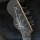 Černá hlava elektrické kytary s ladicími kolíky v černém provedení. Na zadní straně hlavy je bílý nápis "CHRIS CHAPMAN GUITARS" a pod ním bílý podpis…