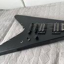 Černá elektrická kytara s tělem ve tvaru V a matným povrchem. Nástroj má dva humbuckery, jeden ovládací knoflík a přepínač zvuků. Na těle není vidné…