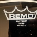 Detailní záběr na černý bubnový kožen. Je na něm bílé logo REMO v podobě koruny a pod ním text PINSTRIPE EBONY MADE IN U.S.A. Povrch je lesklý s vidi…