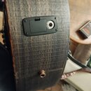 Detail zadní strany akustické kytary s tmavým, strukturovaným povrchem dřeva. Na zadní dece je umístěna černá plastová zásuvka s jedním vstupem pro k…