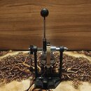 Černý bicí pedál s kovovou konstrukcí a bílým řemínkem. Na horní části je umístěna dlouhá páka s kulatým knoflíkem. Na pedálu nejsou žádné viditelné…