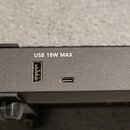 Detail černé kovové části zařízení s dvěma USB porty. Nad porty je bílý nápis USB 18W MAX. Povrch materiálu je matný a texturovaný.