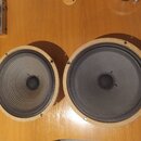 Dvě kulaté reproduktory s tmavě šedými membránami a světle hnědým vnějšíem. Na membránách nejsou viditelné žádné značky ani nápisy. Povrchy jsou v za…