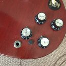 Detail červené kytary s čtyřmi černými oválnými knoflíky. Dva z nich mají nápis VOLUME a dva nápis TONE. V pravé horní části je přepínač s bílým knof…
