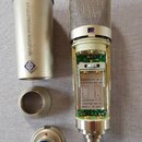 Stříbrný mikrofon s kovovou mřížkou, který je rozložen a odkrutovaný z ochranného pouzdra. Uvnitř mikrofonu je vidět elektronika a štítek s nápisy: E…