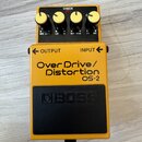 Žlutý kytarový efekt s černým pedalem. Na přední straně je nápis Over Drive/ Distortion OS-2 a na pedalu logo BOSS. Horní panel obsahuje čtyři žluté…