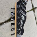 Černá hlava kytary s viditelnou dřevěnou hranou a šesti ladicími kolíky. Na povrchu je bílý nápis CHAPMAN GUITARS a vedle něj velký bílý podpis. Povr…