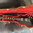 Černá elektrická kytara s tvarem single-cut v hnědém kufru s červeným vnitřkem. Na hlavě je bílé logo Gibson, na hradle jsou dva humbuckery a čtyři o…