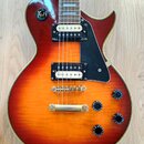 Elektrická kytara s tělem v červeno-oranžovém sunburst provedení a černým plechtrátem. Na horní části těla je vidět kulatý štítek s nápisem „CUSTOM“…