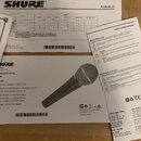 Fotografie zobrazuje dynamický mikrofon značky SHURE v černé barvě s kovovou síťkou. Na těle mikrofonu je viditelné logo SHURE a nápis SM58. Vedle ná…