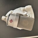 Stříbrný kondenzátorový mikrofon s kovovou síťkovou hlavou, položený na bílé látce. Na těle je vidět logo v červeném kosočtverci a nápis NEUMANN MICR…