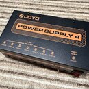 Černý obdélníkový zdroj napájení s oranžovými detaily. Na horní straně je bílé logo JOYO a nápis POWER SUPPLY 4 v oranžovém rámu. Přední strana obsah…