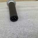 Detailní pohled na spodní část černého mikrofonu s kovovou mřížkou. Je vidět konektor XLR s třemi připájecími piny. Povrch těla mikrofonu je matně če…