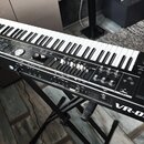 Černý digitální klavír umístěný na černém stojanu. Na přístroji jsou viditelná loga a nápisy Roland a VR-09. Instrument disponuje klávesnicí, displej…