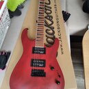 Červená elektrická kytara s tmavým hnedým hmatníkem, položená na kartónové krabici s nápisem „Jackson“ a adresou „WWW.JACKSONGUITARS.COM“. Na hlavě n…