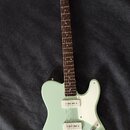 Světle zelená elektrická kytara s bílým pickguardem a hnědým hmatníkem. Na hlavě je viditelné logo Squier. Kytara má dva snímače a tři ovládací knofl…