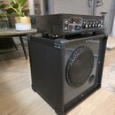 Černý reproduktor v robustním kufrovém provedení s kovovou mřížkou. Na něm je umístěn černý zesilovač s viditelnými elektronkami a ovládacími prvky.…