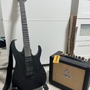 Černá elektrická kytara s matným povrchem a černým popruhkem, umístěná na stojanu. Na hlavě kytary je viditelné logo "Ibanez". Vedle kytary stojí mal…