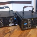 Dvě černé svítící zařízení s rukojitími z kovových profilů. Na zadní stranách jsou viditelné nápisy DMX 512, LASERWORLD a digitální displeje s červen…