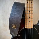 Černý kožený popruh s vyraženým logem a nápisem MUSIC MAN. Popruh je připevněn k basové kytáře s hnědým dřevěným hmatníkem a černým krytem. Povrch ků…