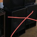 Čtyři černé reproduktorové bedny s kovovými mřížkami. Dvě jsou umístěny v horní řadě a dvě v dolní. Na spodních bednách je červený křížek. Žádné znač…