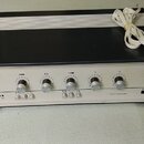 Černý audio zesilovač s bílým předním panelem a bílým napájecím kabelem. Na panelu jsou umístěny pět otáčivých ovratných knoflíků a čtyři malá tlačít…